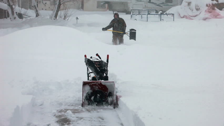 Man walks towards snow blower in deep snow. Deep snow blizzard. Snow blowing in strong wind in sub freezing weather. Winter time hazard and climate nature.