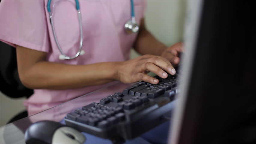 The camera tilts up from a computer keyboard where a pretty young African American nurse or nursing student is working to her face whose expression is that of serious focus on the work she is doing.