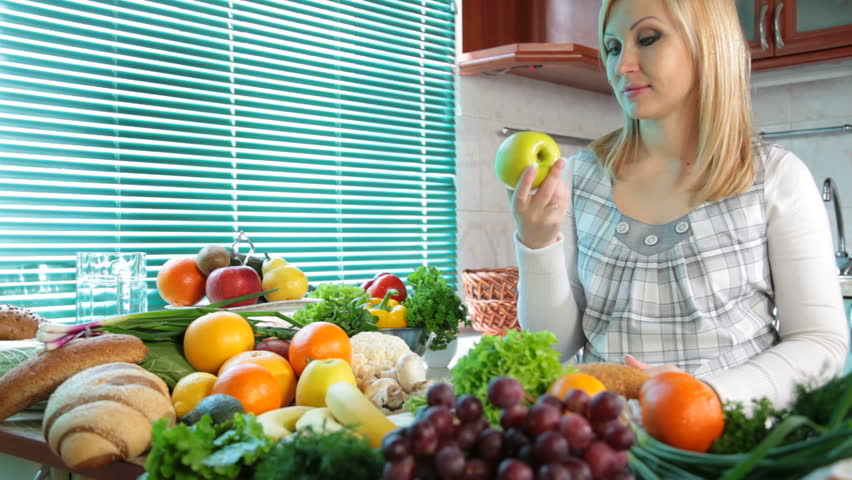 Pregnant woman eating apple near a lot of vegetables and fruits in the kitchen