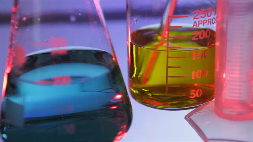 A yellow liquid being stirred in a chemistry beaker with a graduated cylinder and flask containing green liquid in the foreground.