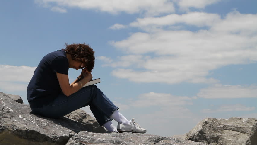 Mature woman holds a bible in her lap with head bowed praying sitting on large rocks with a cloudy blue sky background.