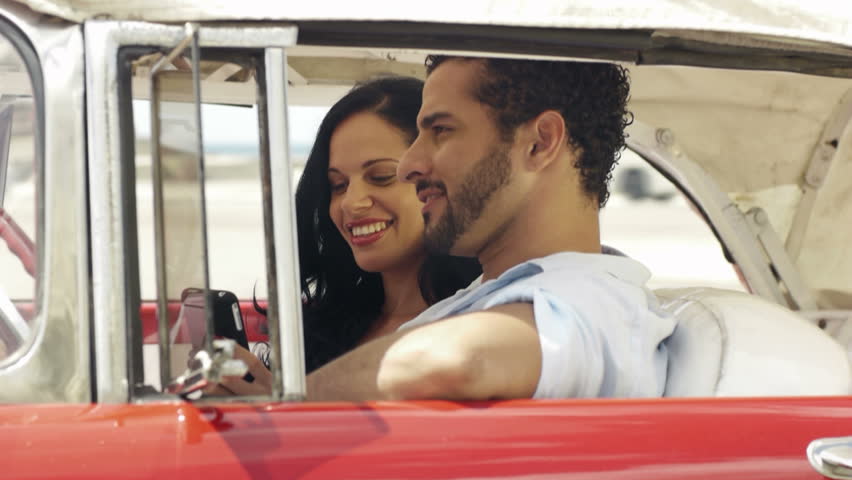 happy young man and woman smiling while taking snapshot with cell phone camera from red vintage convertible car
