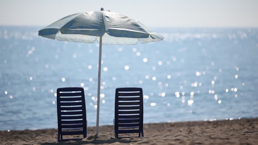 Two empty deckchairs stand on beach under parasol at sunny day