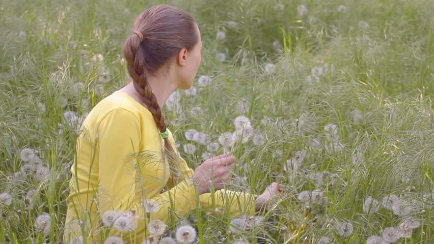 Young woman in yellow blouse collecting dandelions, sitting on weed's meadow, blowing dandelion's seeds, flirting, smiling, throwing them out. 