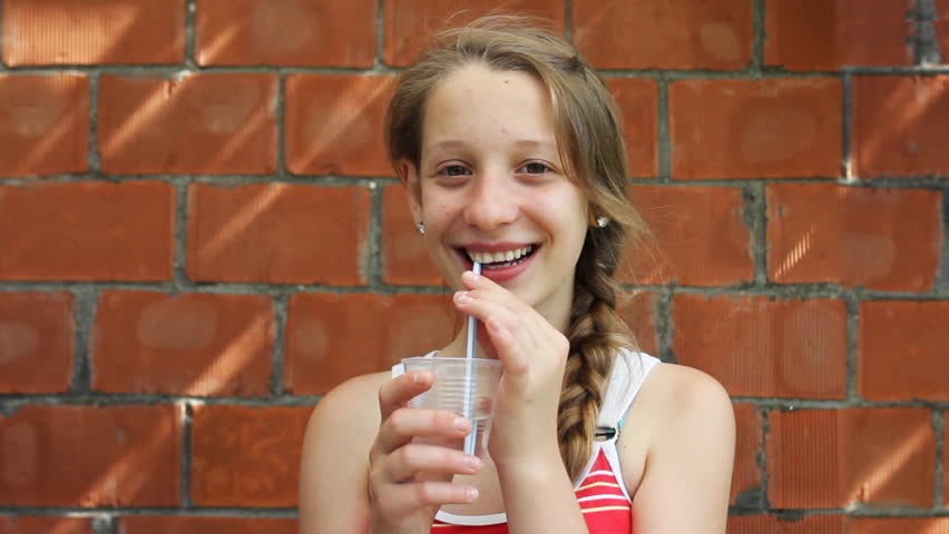 nice young girl sits in front of brick walls and drinks from plastic water cup through straw