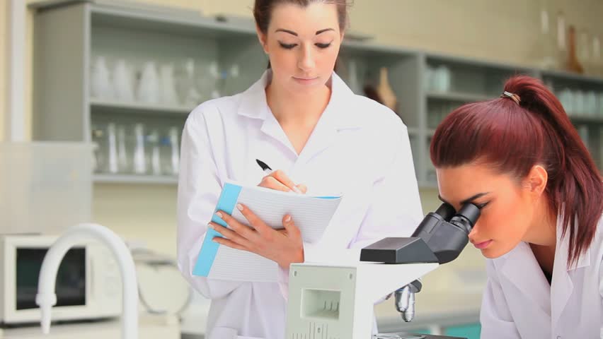 Science student looking in a microscope while her classmate is writing in a laboratory