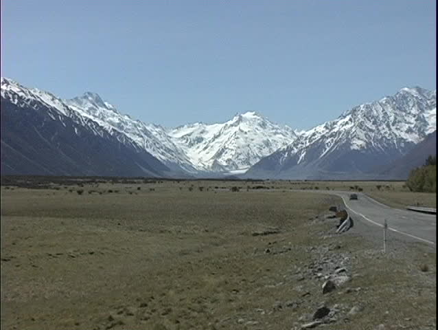 The camera zooms in on snow-capped Mt. Cook in New Zealand as a car approaches from the distance.