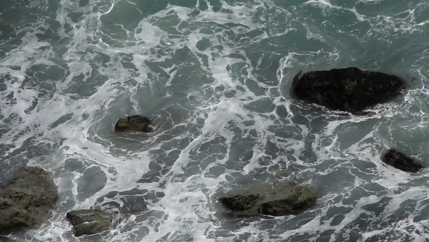 Waves swirl, foam, around boulders.  1080p