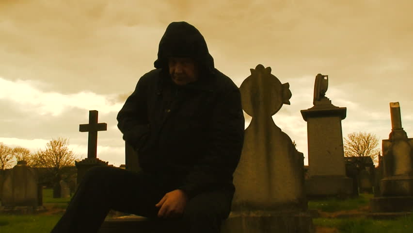  Hooded man sitting among gravestones silhouetted against a stormy sky