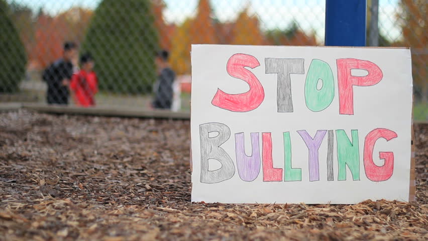 'Stop Bullying' sign. In the background, a group of teenagers push, shove and bully a minority student on the basketball court at school.