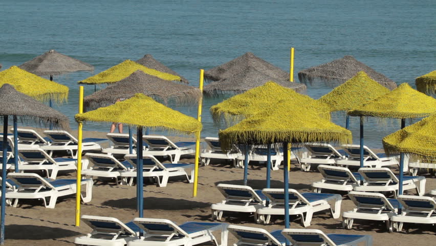 Beach umbrellas on beach of Mediterranean Sea water in background. Vacation and tourist destination for summer fun and enjoyment. Bright yellow with white and blue beach lounge chairs.