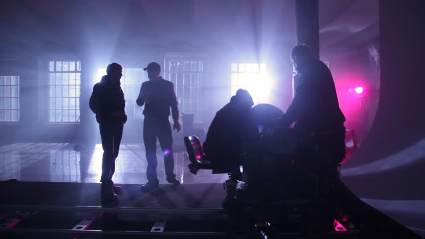 film crew adjusts equipment on rail cart in dark room shined artificial light behind windows