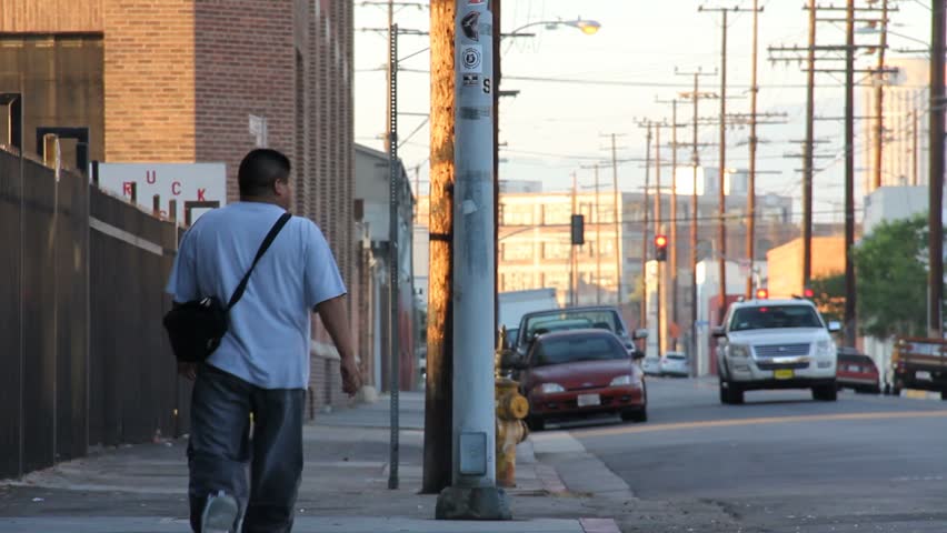 Two pedestrians walk down a sidewalk on the left side. Traffic and telephone poles are also in frame.