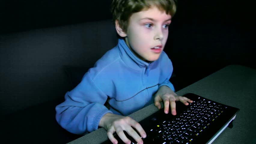 Little boy plays video game on big screen with keyboard and chews gum in dark room