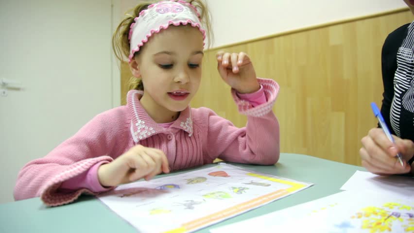 Little girl sits at table with book and utters words, teacher writes near her