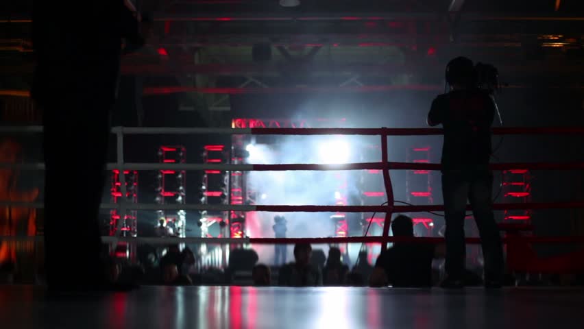 Fighter comes from metal gate with illumination, peoples silhouettes, unfocused view from boxing ring