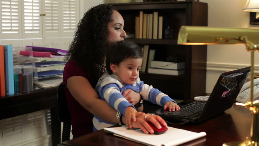 A pretty Hispanic mom holds her son while working on a laptop in her home office.