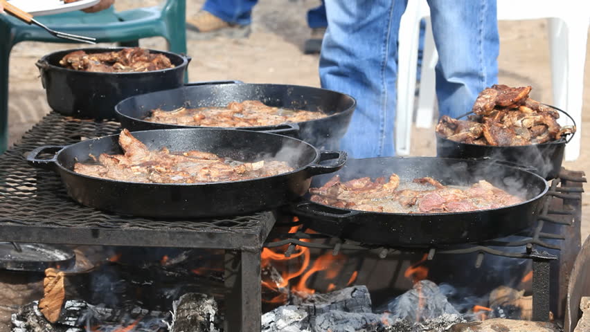 Lamb steaks and chops cooking in caste iron pans over an open outdoor fire. Men turning meat.  Cowboy and sheep camp bbq and party. Fresh meat with smoke and oil boiling near Dutch Oven