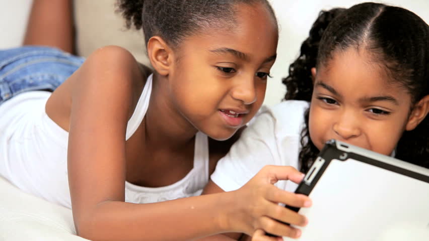 Cute little african american girls lying on their home sofa using a wireless tablet