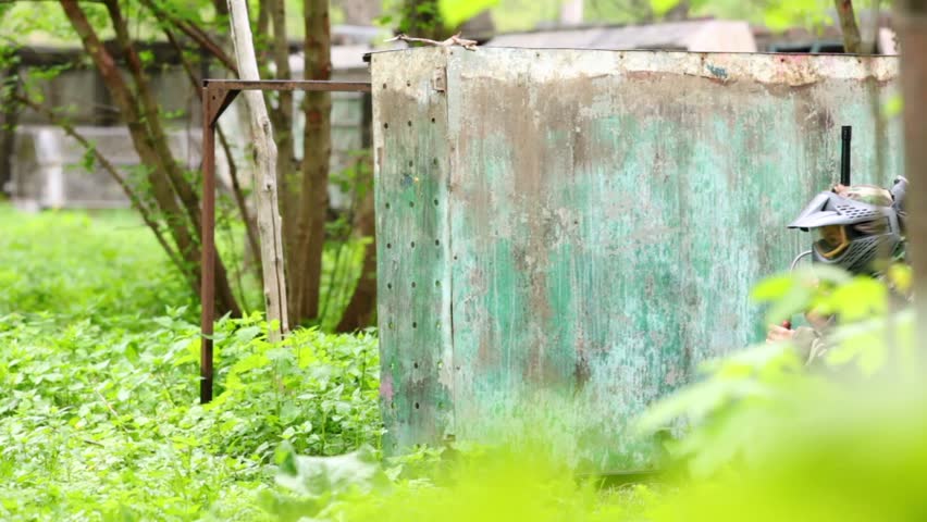 Boy paintball player sits in ambush behind metal fence, then stands up and runs away