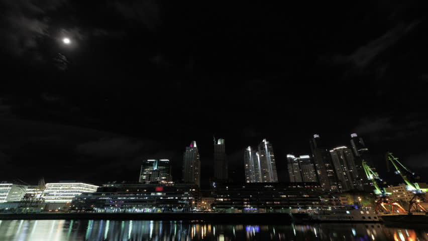 Clouds fly over modern skyscrapers and the moon at night. Photo time-lapse with ultra wide-angle across a harbor dock  (Puerto Madero, Buenos Aires, Arg.; Mar 2012; DSLR).
