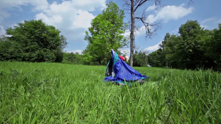 Little boy pitches tent on grass glade under blue sky with clouds