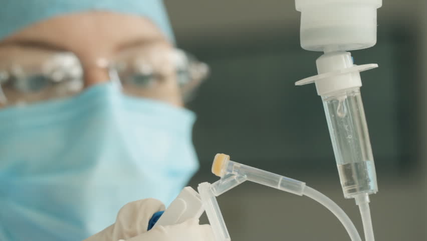 Adjusting Medical IV Drop Rate (HD). Nurse controlling intravenous drip speed rate showing control wheel. Nurse is out of focus, drip IV & hand in focus.