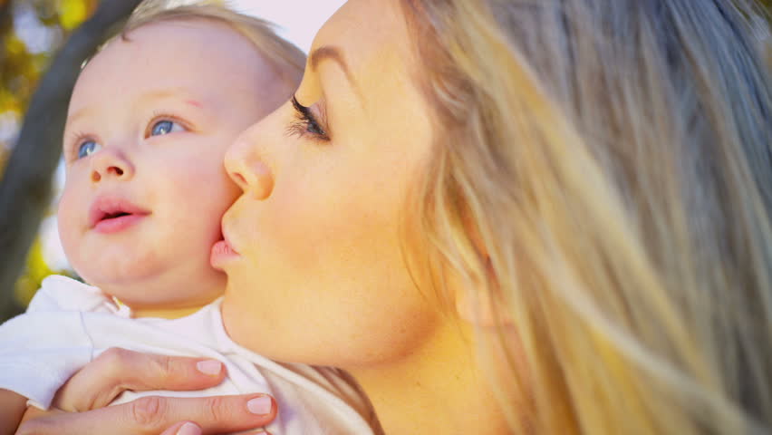 Portrait in close up of young blonde mother outdoors giving her smiling baby a kiss