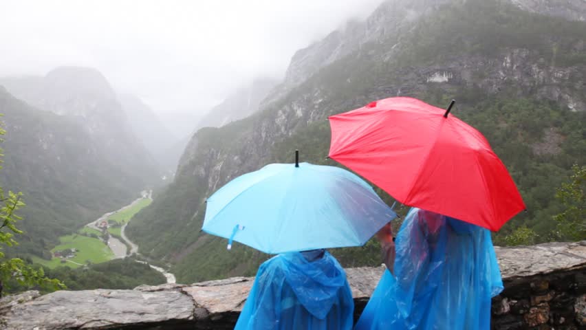 mother and son in raincoats and under umbrellas look valley in woody mountains