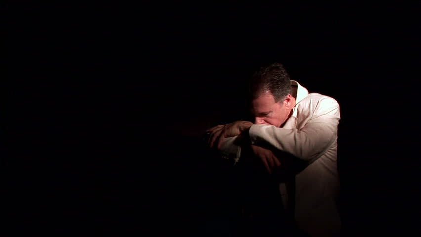 A man, who appears to be very troubled, sitting astride a bent back chair in a dark room