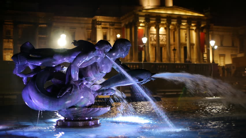 One of the fountains in Trafalgar Square in London. Night, lit by underwater lights. National Gallery entrance in the background.