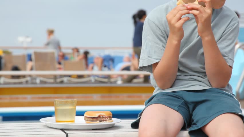 boy sits near plate with hamburger, eats and drinks, and behind people have rest in chaise lounges