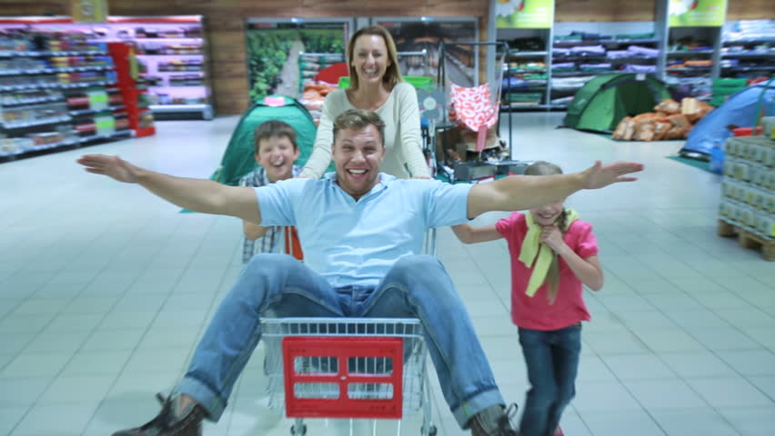 Mom and two kids having fun wheeling daddy around store in the market shopping cart.