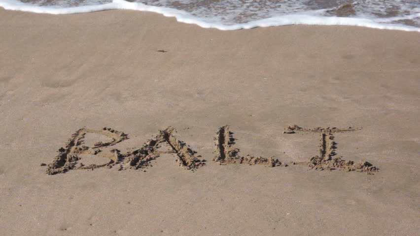 The word of the popular vacation destination Bali, Indonesia written in the sand with a wave coming up on the shore next to it.