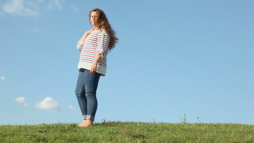 Young girl bents backward on grass hill at sunny summer day
