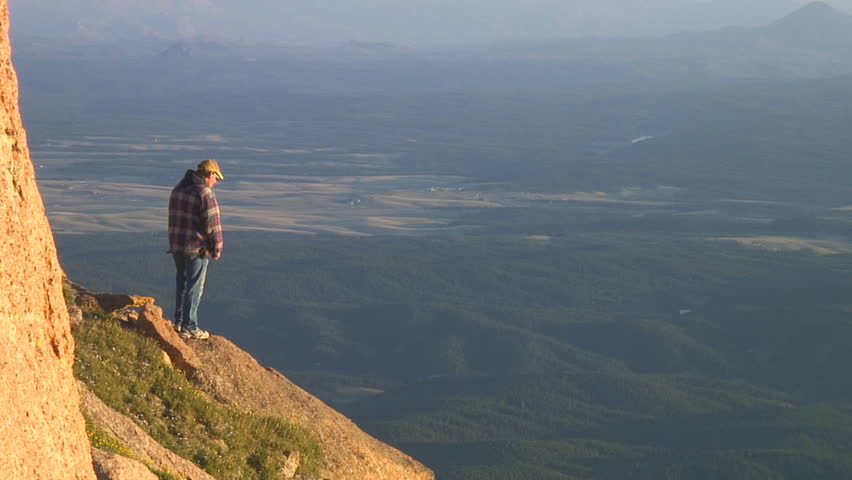 Man stands on mountain cliff in morning sunlight pointing out the view below