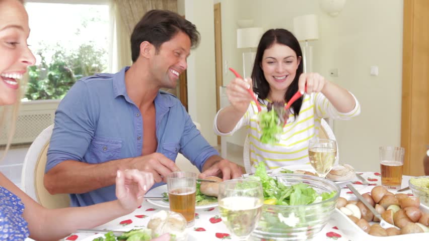 Couples seated around dining table serving one another before picking up glasses for toast.
