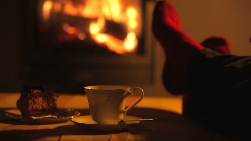 Man relaxes by warm fire then stirs drink with teaspoon before taking sip and returning to saucer.