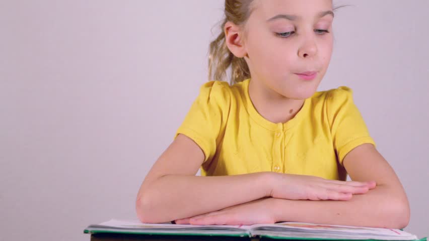 Little girl in yellow shirt reads book aloud and turns page isolated at white background
