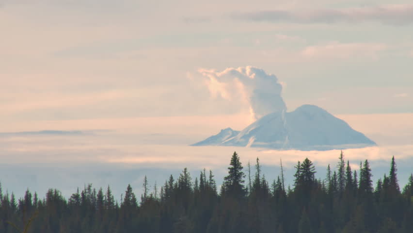 A timelapse of a conical volcano in Alaska (Mt. Redoubt) spewing steam with low clouds passing by.