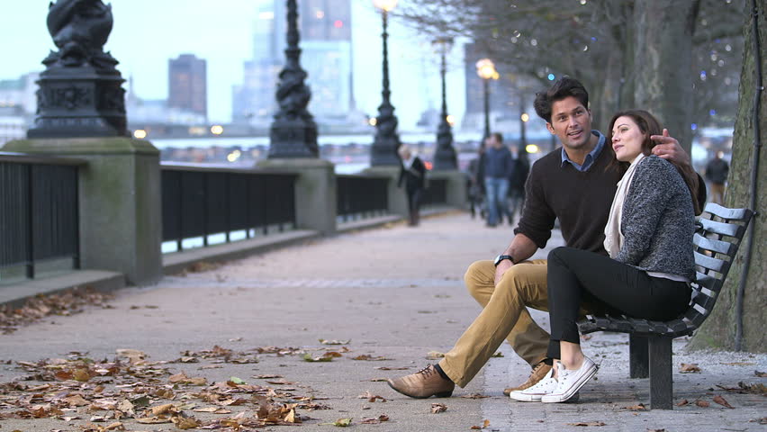Casually dressed couple enjoy a romantic moment together, sitting on a London bench beside the river thames. The light of the day is just beginning to fade