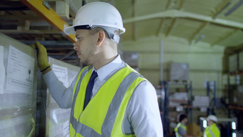 A worker in a warehouse climbs a ladder to check stock on a high shelf and he turns and smiles to the camera. 2 of his colleagues can be seen talking in the background.