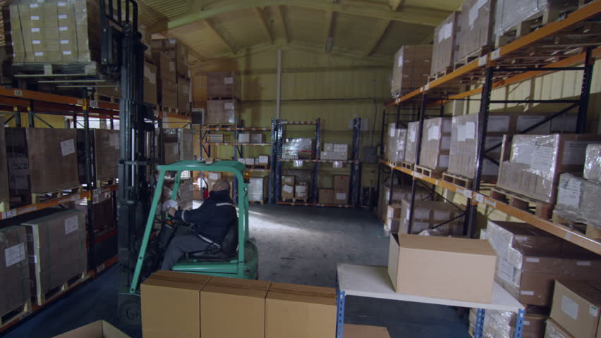 A worker in a warehouse is using a forklift truck to move stacks of boxed goods around