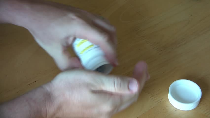 Shaky hands open pill bottle, and then pour medicine onto wood table before putting top back on.