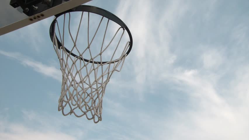 Clip of businessman in tie attempts to dunk a basketball, shot from underneath rim and net during afternoon with sky as background..