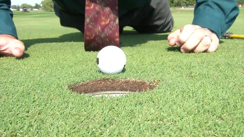 Businessman blows golf ball into hole on green after a couple of attempts, pulls ball out, gets up, and replaces yellow pin, in sunlight.
