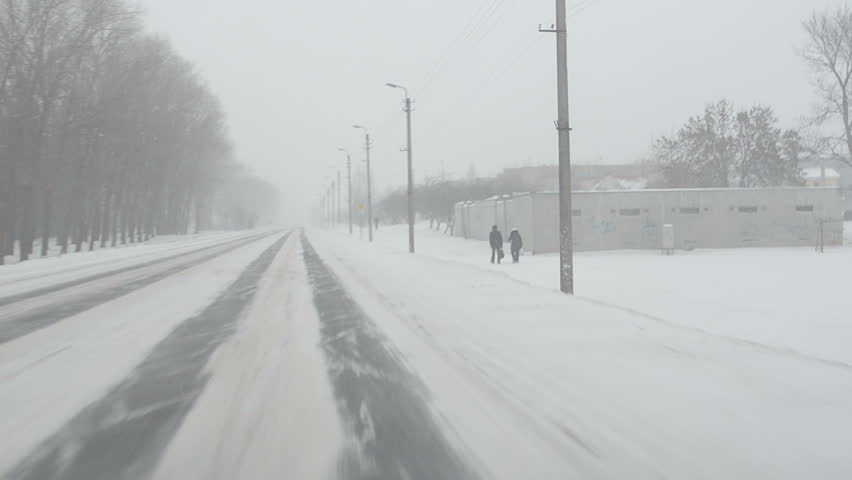 car go on road near small town and snowstorm snow falling. people walk. front car window view.