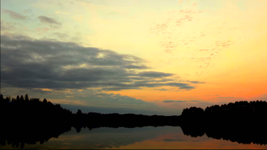 Time Lapse: Sunset clouds glow orange and purple above a tranquil lake in the Wisconsin North Woods. Slow zoom in.