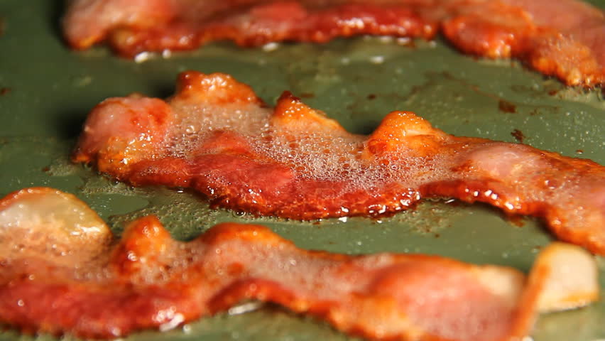 Bacon 4. Three strips of bacon frying in a pan turned over with tongs. Later in the cooking process.