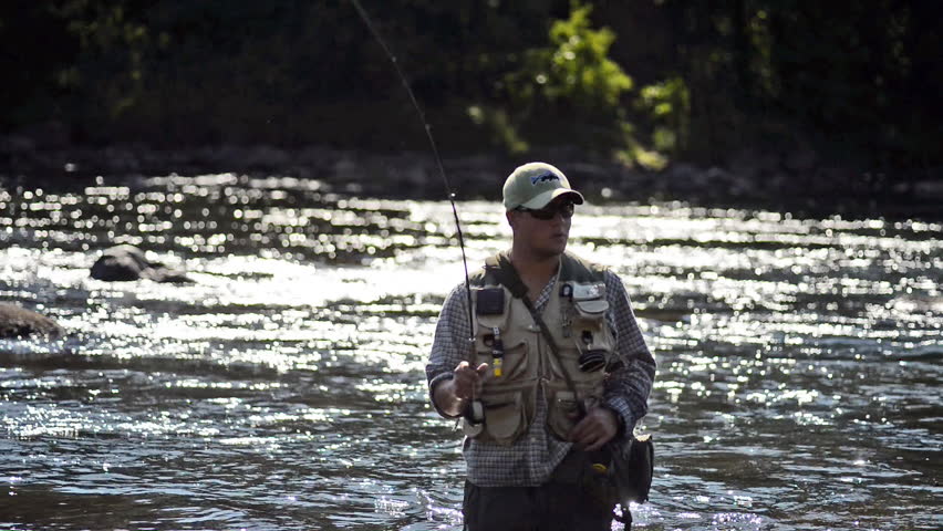 A man in waterproof overalls stands in a river and casts his lure with his rod while he spends his day fly fishing.
